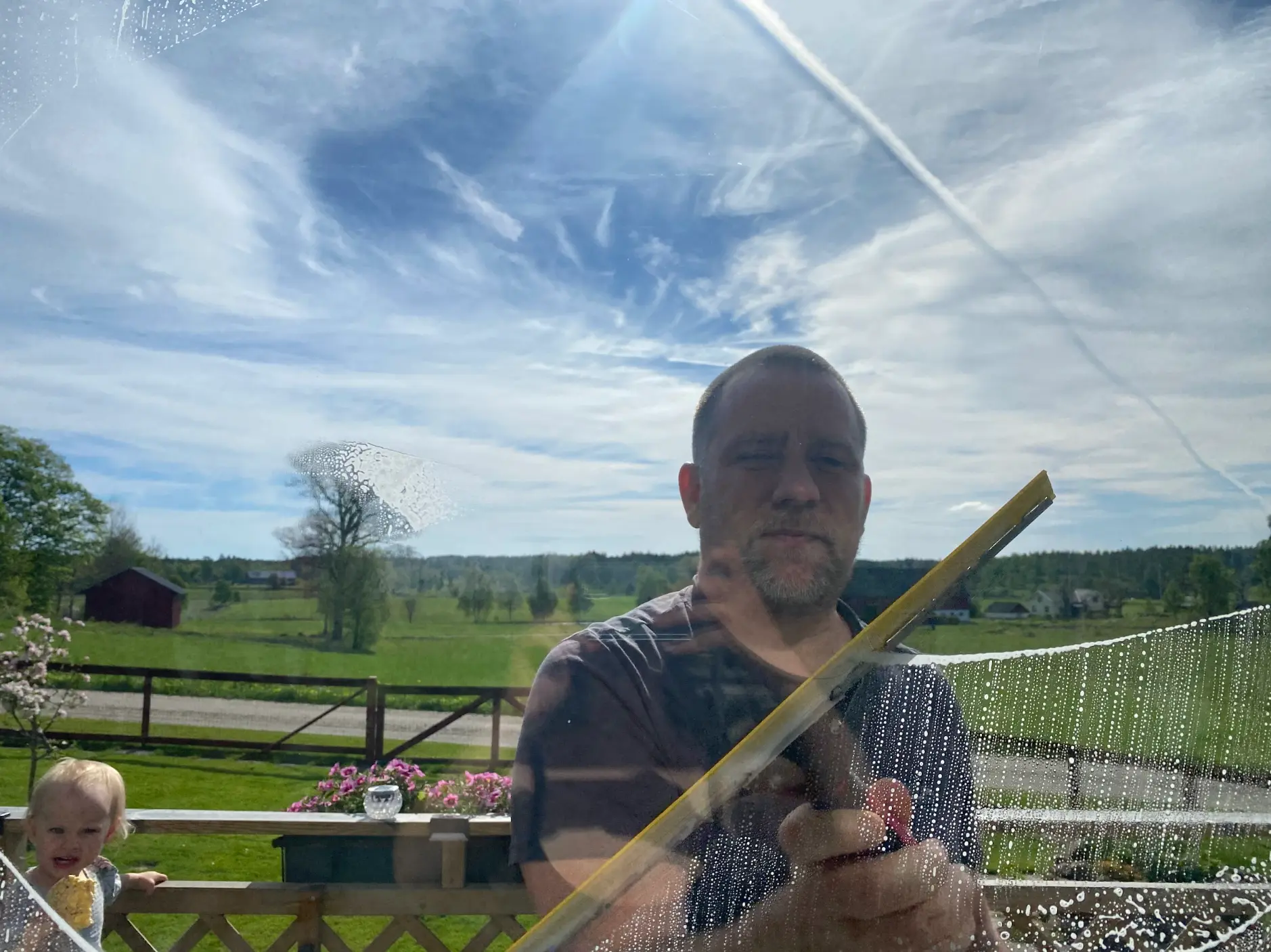 Kristopher putsar ett fönster med skrapa, med utsikt över landskapet utanför Ulricehamn